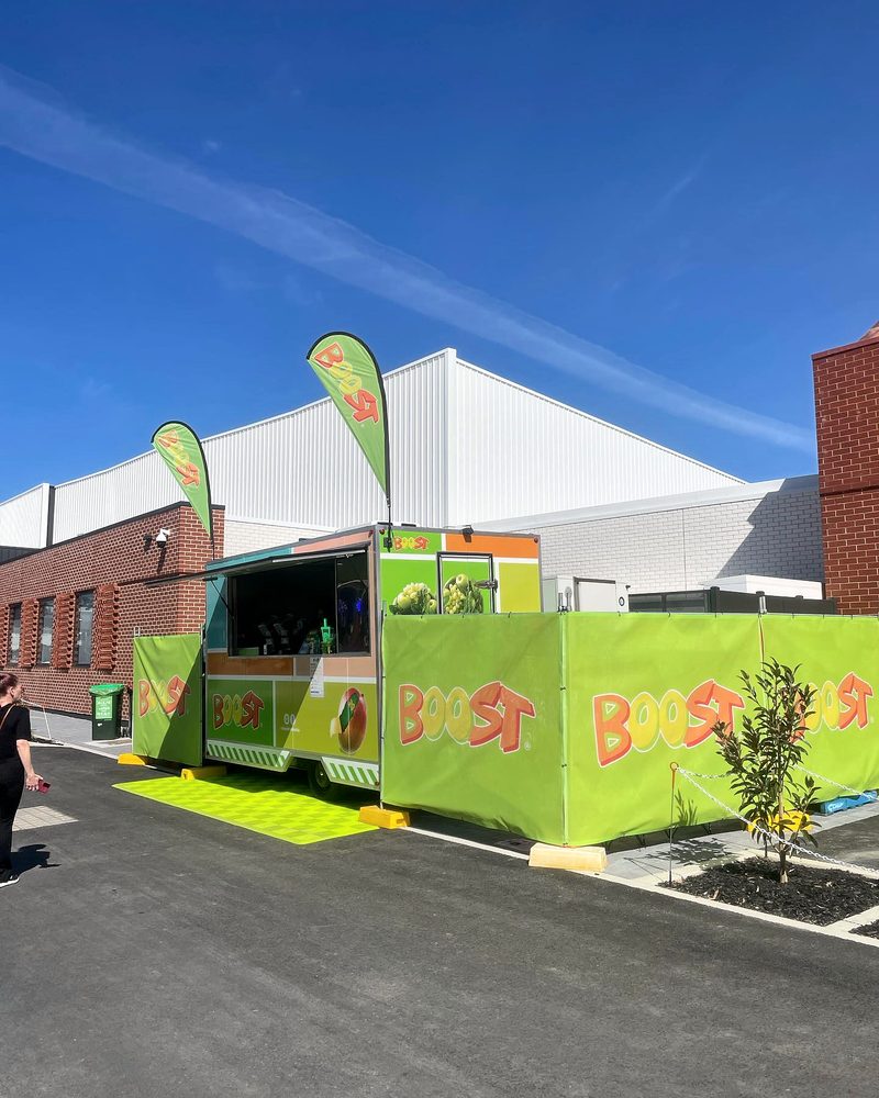 The Boost Juice trailer at a second spot, an ULTRAGRID Electric Green mat laid beside it and green Boost screens along the front