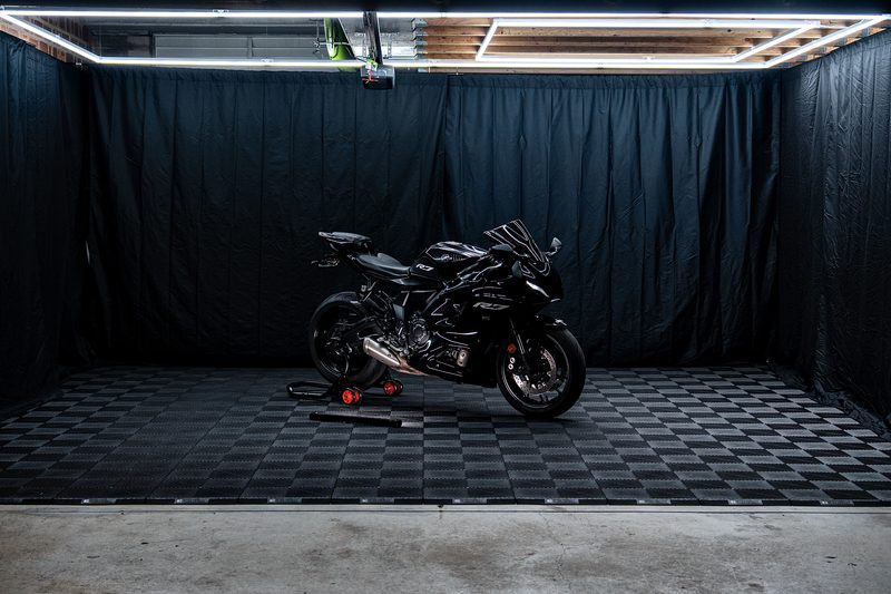 A black sportbike on a paddock stand in the middle of a Jet Black ULTRAGRID floor, black curtains behind, a rectangle of ULTRABEAM tube lights along the ceiling