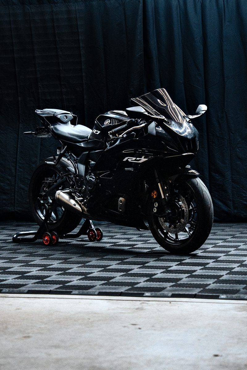 A black sportbike with R7 on the fairing on a red paddock stand on the Jet Black ULTRAGRID floor, concrete threshold at the door, black curtains behind