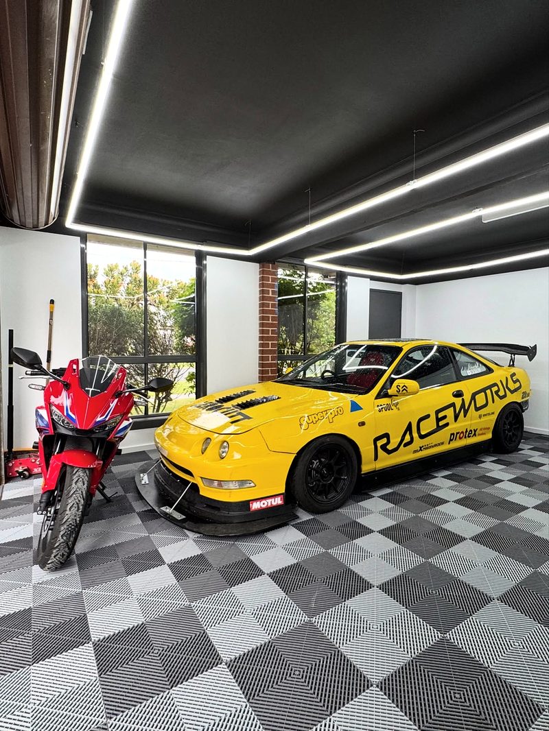 A yellow Integra race car in Raceworks livery and a red Honda CBR on a chequer floor of ULTRAGRID Silver Grey and Space Grey, white walls, dark ceiling with rectangles of ULTRABEAM light